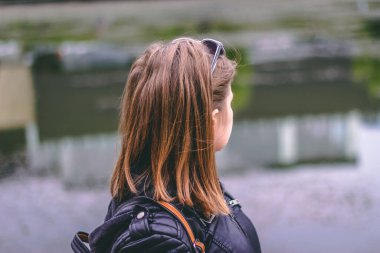 Kısa kahverengi saçlı genç bir kadının gölde düşünceli bakışları. Deri ceketli ve güneş gözlüklü bir kız doğada hayal kuruyor.