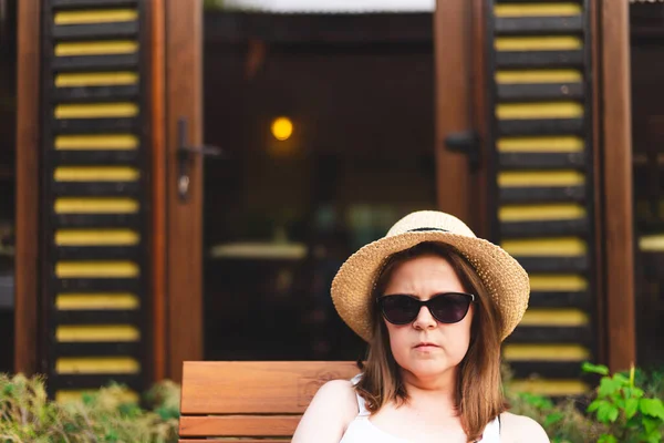 Young relaxed woman wearing sunglasses and a yellow straw hat sitting in a wooden chair at a bar outdoor. Pretty girl with a casual summer outfit enjoying summertime at a patio