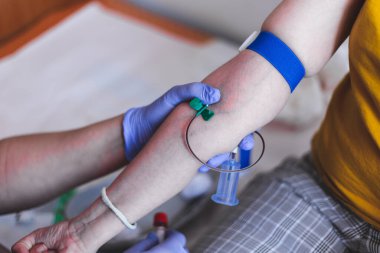 Kadın hemşire, hastanede mavi turnikeli bir genç kızdan plastik şişede kan alıyor. Kadın doktor hasta elinden küçük bir iğneyle sıvı örnekleri alıyor.