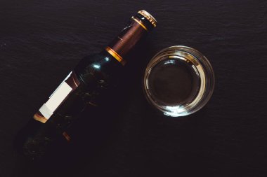 Siyah masada bir şişe ve bir bardak bira. Yanında bardak olan işlenmemiş kahverengi bir içki. Cam alıcılarında soğuk ve ferahlatıcı bir içecek.