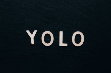 Yolo kelimesi arka plan olarak siyah bir masanın ortasına yerleştirilmiş ahşap harflerle yazılmış. Koyu renkli bir yazı tahtasındaki klasik yazı tipi ile motive edici bir kelime
