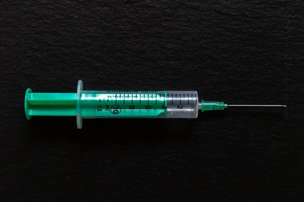Green and transparent syringe with needle placed on black background in a horizontal position. Medical tool used for injecting liquid substances. Concept image for health care and addiction