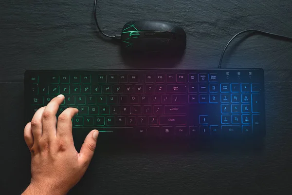 One man hand on rgb computer keyboard and mouse placed next to it on black background. Colorful modern devices for gaming on stone textured desk