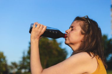 Sıcak bir yaz gününde arka planda mavi gökyüzü olan plastik şişeden soda içen genç bir kadın. Yazlık kıyafetli güzel kız doğada dinlenirken siyah içeceğin tadını çıkarıyor.