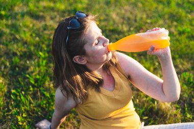Dışarıda güneşin altında oturmuş portakal suyu içen genç bir kadın. Sıradan giyinmiş bir kız kendini iyi hissediyor ve yaz günü soğuk, ferahlatıcı organik içeceklerin tadını çıkarıyor.