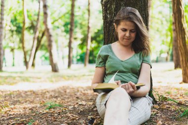 Ağaca yaslanırken dışarıda kitap okuyan gülümseyen kadın. Sıradan giyinmiş bir kız elinde kitap tutarken parkta dinleniyor. Doğada rahatlamanın basit bir yolu.