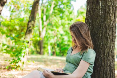 Genç bir kadının parkta bir ağaç gövdesine yaslanırken kitap okurken çekilmiş bir fotoğrafı. Kahverengi saçlı sıradan giyinmiş kız öğrenci yaz günü dışarıda öğreniyor.