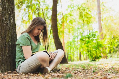 Doğanın dışında okuyan bir kadın. Öğrenci yerde düşen yaprakların üzerinde otururken dışarıda çalışıyor.