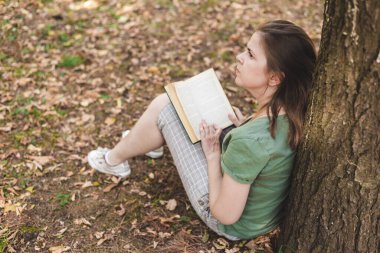 Dışarıda kitap okuyan bir kadın. Öğrenci kampüste iyi vakit geçiriyor. Ağaç Burk 'e yaslanıp kafasını kaldırıyor.