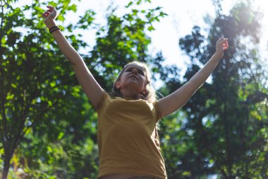 Bir yaz günü zafer işareti olarak iki elini de havada tutan güzel genç bir kadın. Güneşli bir günde doğada mutluluk ve neşe ifade eden sıradan giyinmiş bir kız. Başarı için resmi kavra