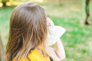 Dışarıdaki beyaz bir peçeteye burnunu silen genç bir kadının arka görüntüsü. Kahverengi saçlı kız parkta yürürken sezonluk alerji belirtileri gösteriyor.