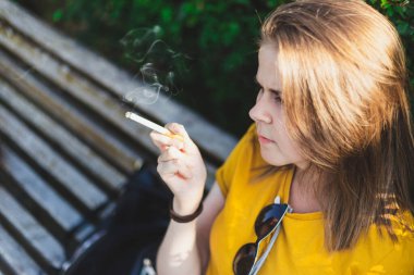 Güneşli bir günde dışarıda sigara içen genç bir kadının yan görüntüsü. Kahverengi saçlı güzel genç kız parkta bir bankta dinlenirken elinde puro tutuyordu. Toksik ve tehlikeli alışkanlıklar insanlar tarafından geliştirildi.