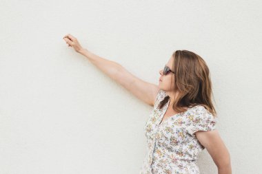 Bir kadın eline fotokopi aletiyle uzanırken beyaz duvara yaslanıyor. Beyaz binanın yanında duran gömlek ve güneş gözlüğü takan genç bir kadın. Milenyum kızı açık hava modası