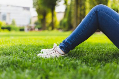 Parktaki yeşil çimlerin üzerinde oturan kadın tuvalden ayakkabılar ve kot pantolon giyer. Sıradan giyimli, beyaz spor ayakkabılı genç bir kız. Modern ve şehirli kadın ayak giyimi yaz günlerinde doğada