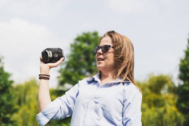 Mutlu, güzel genç bir kadın bir yaz günü dışarıda küçük bir hoparlör tutuyor. Kahverengi saçlı, gülümseyen, güneş gözlüklü, parkta müzik dinlerken şarkı söyleyen tatlı bir kız.