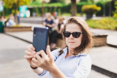 Doğada telefonla fotoğraf çeken genç bir kadın. Sıradan giyinmiş gözlüklü bir kız küçük cep telefonuyla fotoğraf çekiyor. Kadın dışarıda pratik bir aktivite yaparak eğleniyor.