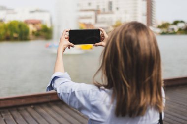 Göl manzarasına cep telefonuyla fotoğraf çeken genç bir kadın. Kahverengi saçlı, mavi gömlekli, doğa resimleri yapan güzel bir kız. Rahatlatıcı ve eğlenceli etkinlik