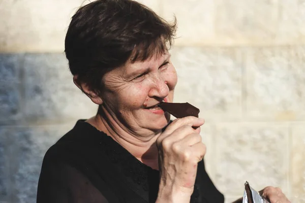 Dışarıda çikolata yiyen son sınıf güzeli bir kadın. Tatlı şekerlemelerin tadını çıkarırken kendini iyi hisseden mutlu tatlı yaşlı kadın.
