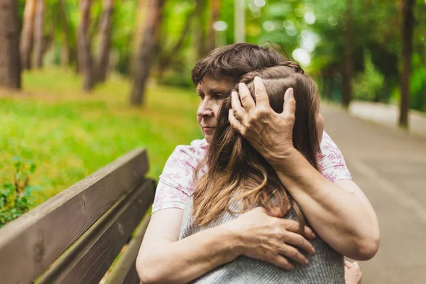 Happy mother holding daughters head with one hand while sitting on a bench in the park. Cute and kind woman showing love and affection to young person with brown hair outdoors