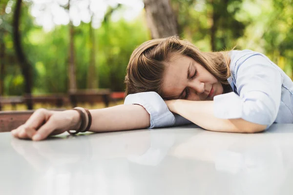 Tired young woman sleeping on one hand on a table outside. Beautiful girl with brown hair taking a nap in nature. Exhausted female worker relaxing in the park with her eyes closed