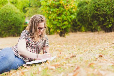 Dışarıda yazan kadın. Kolej kampüsünün dışında okuyan öğrenci gözlük takıp çimlerin üzerinde yatıyor.