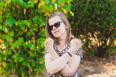 Güçlü genç bir kadın açık havada savunma işareti olarak yumruğunu havada sıkıca tutuyor. Kahverengi saçlı, güneş gözlüklü, güler yüzlü kız parkta el hareketi yapıyor.