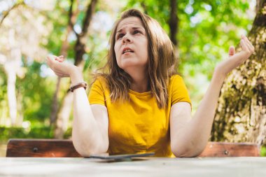 Kafası karışmış bir kadın dışarıda bir masada otururken yukarı bakıyor. Belirsiz bir ifadeyle omuzlarını kaldıran kafası karışmış bir kız.