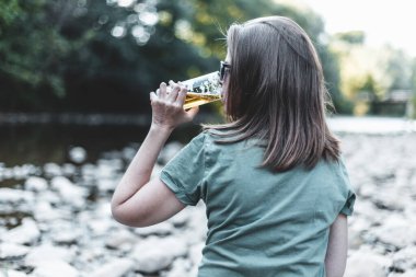 Dışarıda soğuk bira içen mutlu güzel kadın. Güneşli bir günde, ormanın doğal ortamında yaz içkisinin tadını çıkaran rahat bir kadın.