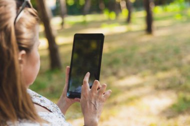 Dışarıda tablet kullanan genç bir kadının dikiz görüntüsü. Kahverengi saçlı kız parkta ekrana dokunurken mobil bir cihaz tutuyor.