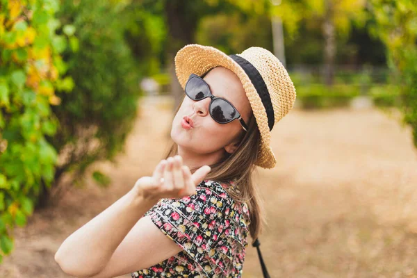 Young woman sending air kiss outdoors. Beautiful girl wearing summer hat and sunglasses blowing kisses. Trendy millennial relaxing and having fun in nature