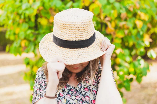Shy woman covering her face with a summer hat outdoors. Pretty girl feeling humiliated while hiding behind straw hat in the park. Cute and adorable gesture made by young female in sign of shame