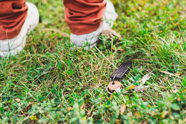 屋外の芝生の中で失われたキーのペア 公園の金属ドアの開口部の横に緑の草原に立って白いスニーカーを持つ人のクローズアップショット — ストック写真