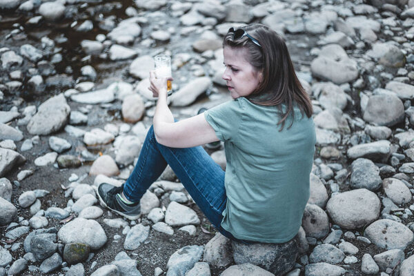 Woman with beer sitting on rocks at a river's edge. Lonely traveler enjoying the moment while holding a glass of alcohol