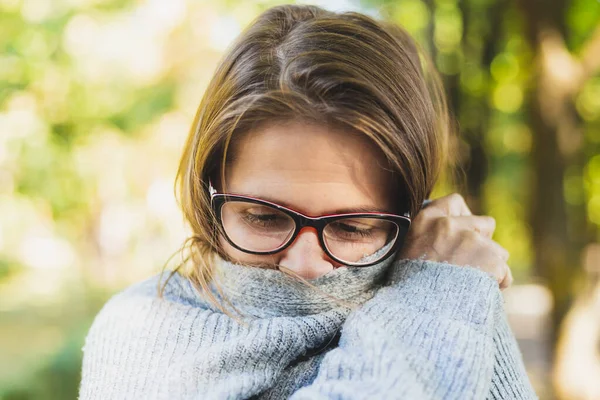 Infected woman with cold virus covering her mouth outside. Pretty girl wearing eyeglasses holding hands over her face while having flu symptoms. Young female suffering from fever. Concept image for epidemic