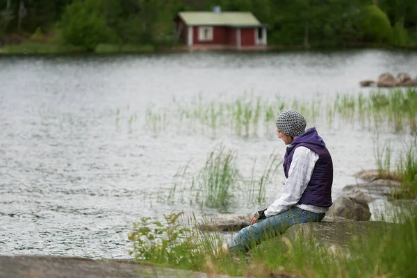 Young woman wearing hat and purple waistcoat relaxing on lakeside in breezy summer day.