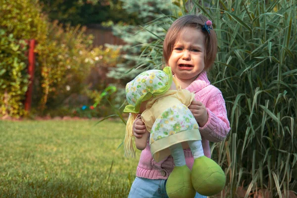 Una niña pequeña con una muñeca en la mano, de pie junto a la hierba alta y llorando . — Foto de Stock