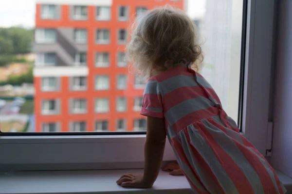 Pequeña chica rizada con el pelo rubio mirando hacia fuera desde la ventana en el barrio de varios pisos — Foto de Stock