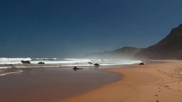 Ocean coast with footprints on sandy beach, black rocks, blue sky and huge waves