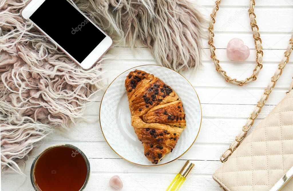 Morning flatlay with croissant and tea, smartphone, handbag on white wooden background. Women's breakfast. Concept of wellbeing, inner peace and simple pleasures.
