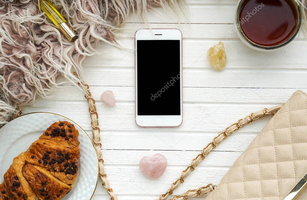 Morning flatlay with croissant and tea, smartphone, handbag on white wooden background. Women's breakfast. Concept of wellbeing, inner peace and simple pleasures.