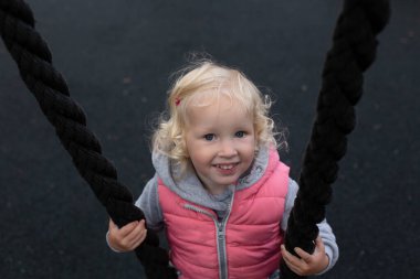 Sarı kıvırcık saçlı, büyük mavi gözlü, siyah ipten yapılma salıncakta sallanan neşeli kız.