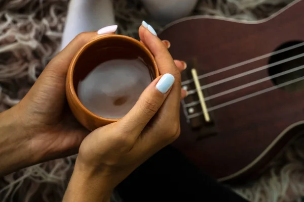 Cozy photo of womans hands holding cup of tea with ukulele on backdrop. Fall or winter time concept. — Stock Photo, Image