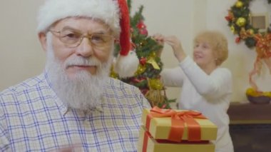 Yaşlı adam karısını Noel hediyesi ile sürpriz yapmak istiyorum. Kadın arka plan, Noel ağacı süsleyin. Olgun adam Santas şapka Haritayı işareti sessizlik kamera takıyor. Adam ve iki kadın geldi