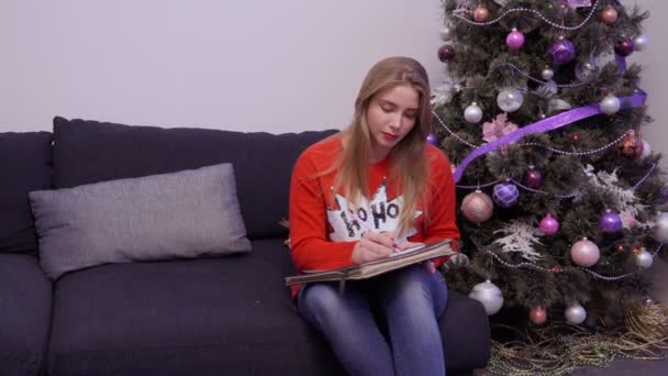 Молодая девушка пишет письмо сидя на диване рядом с елкой — стоковое видео