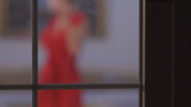 Kamera çekimde. Pencerenin dans kırmızı elbiseli güzel bir kadın. Odak bulanık. Bir pencereden bakıyor.
