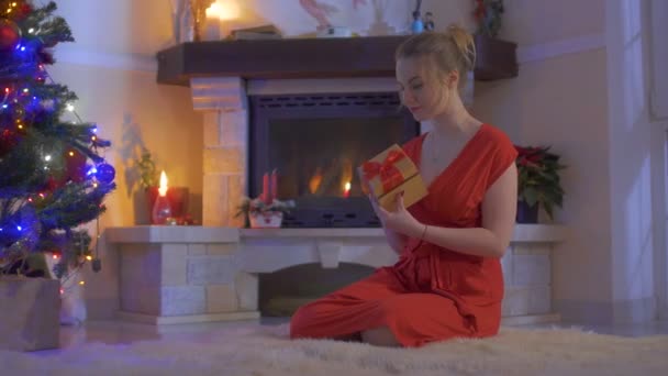 Молодая женщина сидит на сказочном ковре и открывает свой подарок возле елки — стоковое видео