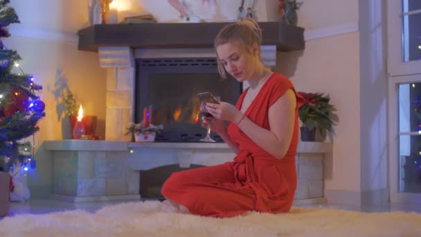 穿着红色连衣裙的年轻女孩坐在圣诞树旁的地板上, 用她的电话 — 图库视频影像