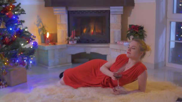 Beautiful woman in red dress drink wine and lies on carpet near Christmas tree