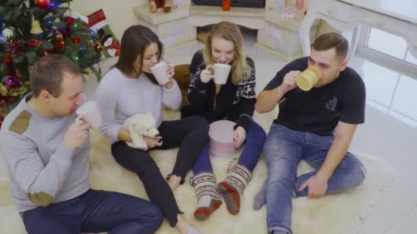 Czterech młodych ludzi pije kawę lub herbatę, siedząc na podłodze w pobliżu choinki — Wideo stockowe