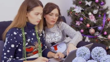 İki genç kız arkadaşım kazak oturur evde kanepe ve internet deposunda online Noel alışveriş yapar. Çekici kızlar birlikte evde rahatlatıcı ve tatil için hazırlar.